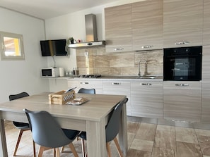 A modern kitchen with light wood cabinets, a stainless steel range hood, a black oven, and a dining area with a wooden table and grey chairs.
