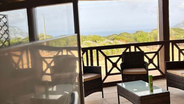 A covered outdoor seating area with wicker furniture and a glass table, overlooking a mountainous landscape. - MATTEOTTO RESIDENCE, 2-BEDROOM APARTMENT, (Sardegna)