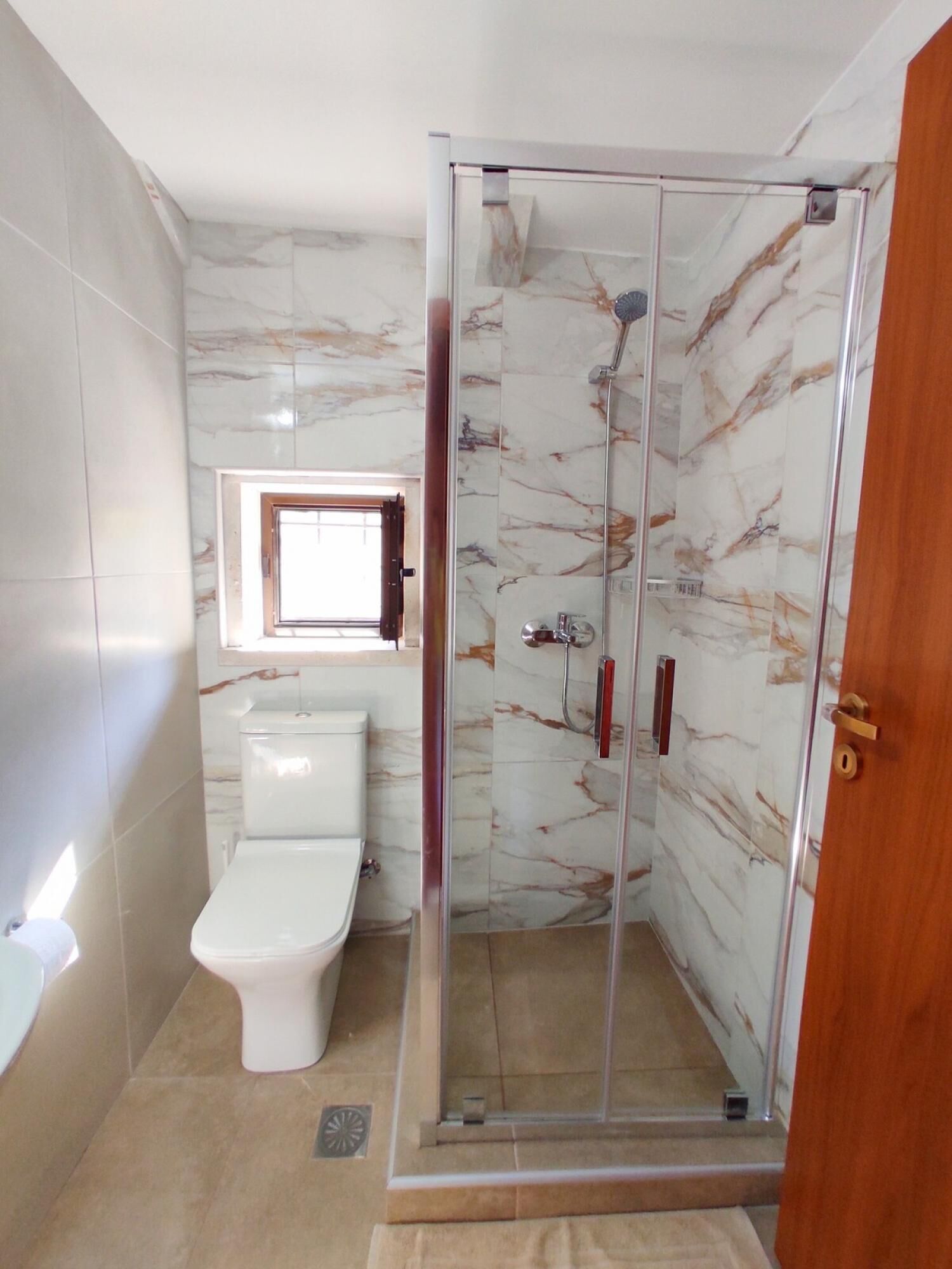 A bathroom with a glass-enclosed shower, a toilet, and a window above the toilet.