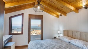 A bedroom with a wooden ceiling, a bed with white bedding, a nightstand, a lamp, and a view of the sea through a window.