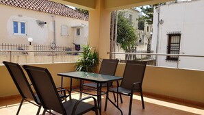 A balcony with a glass table and four chairs, overlooking a street with buildings and a fence. - Ilia s Apt in Chania (Chania)