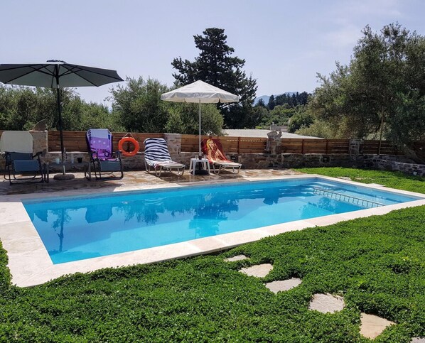 A swimming pool with lounge chairs and umbrellas, surrounded by greenery and a stone wall. - Traditional Villa in Fres (Apokoronas)