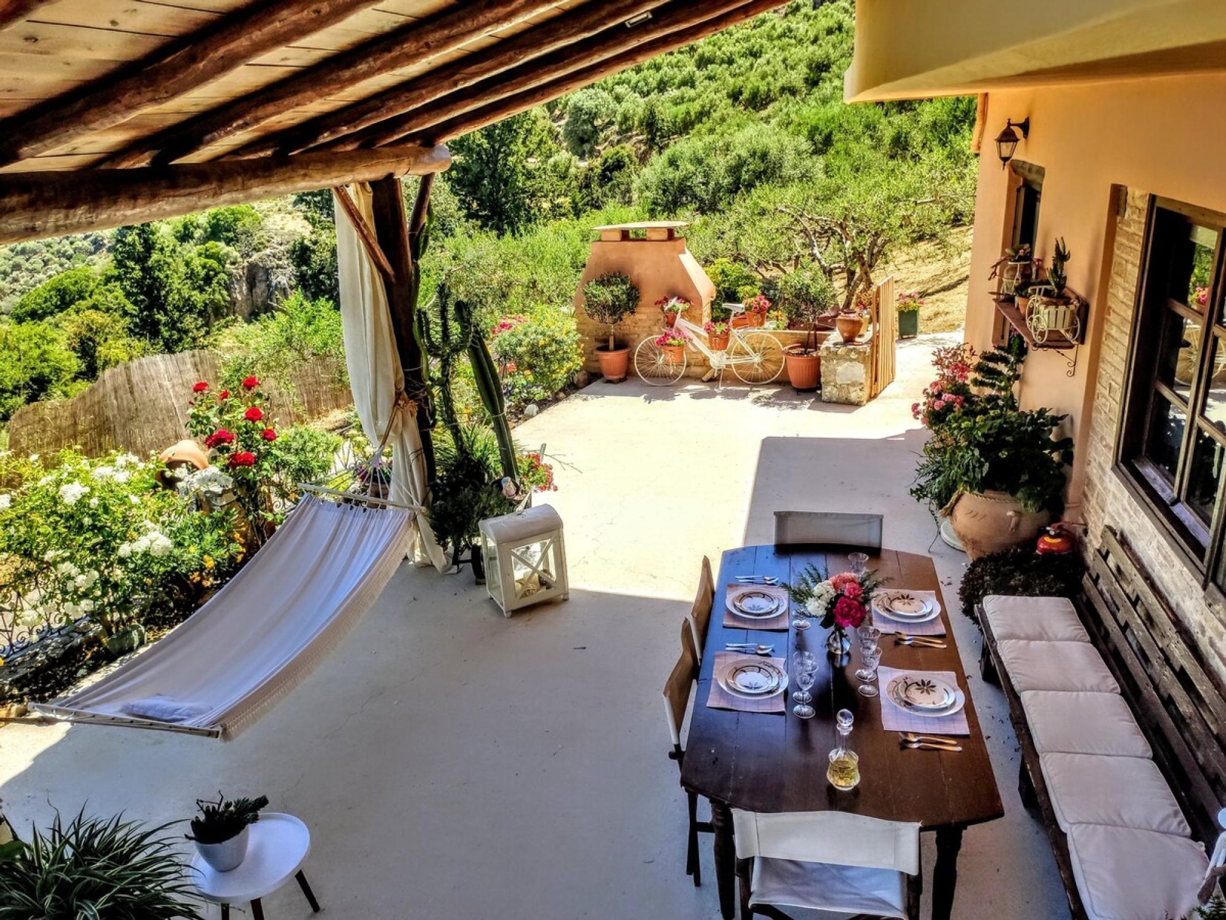 A covered outdoor dining area with a table set for two, surrounded by potted plants and a hammock.