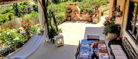 A covered outdoor dining area with a table set for two, surrounded by potted plants and a hammock.