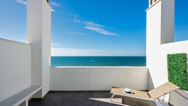 Varanda com vista para o mar, uma parede branca e uma pequena mesa com uma cadeira.