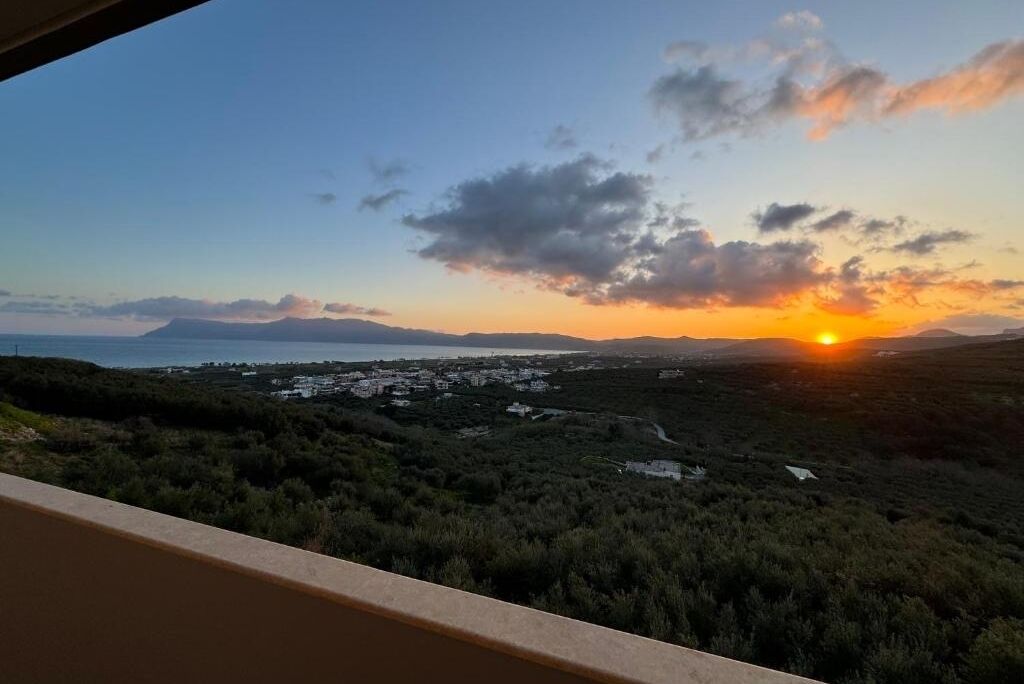 Un paisaje costero con atardecer, vegetación y edificios a lo lejos.