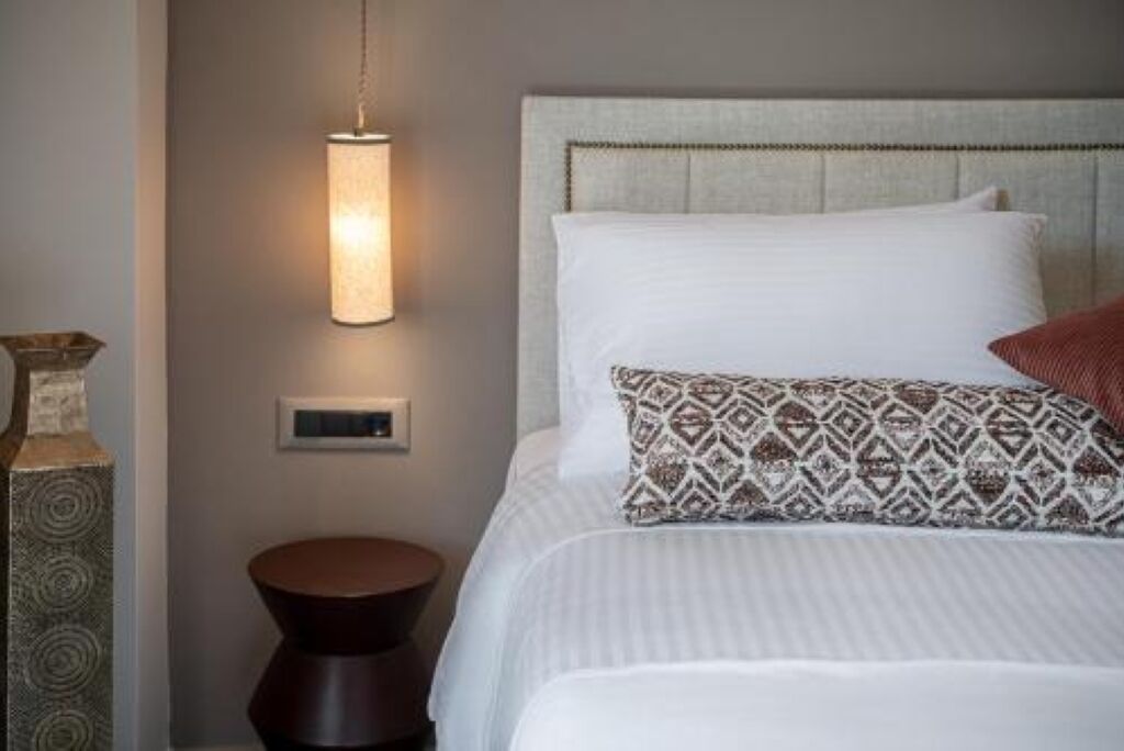 A neatly made bed with a patterned pillow, a round wooden nightstand, and a wall-mounted light fixture.
