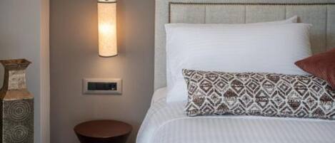 A neatly made bed with a patterned pillow, a round wooden nightstand, and a wall-mounted light fixture.