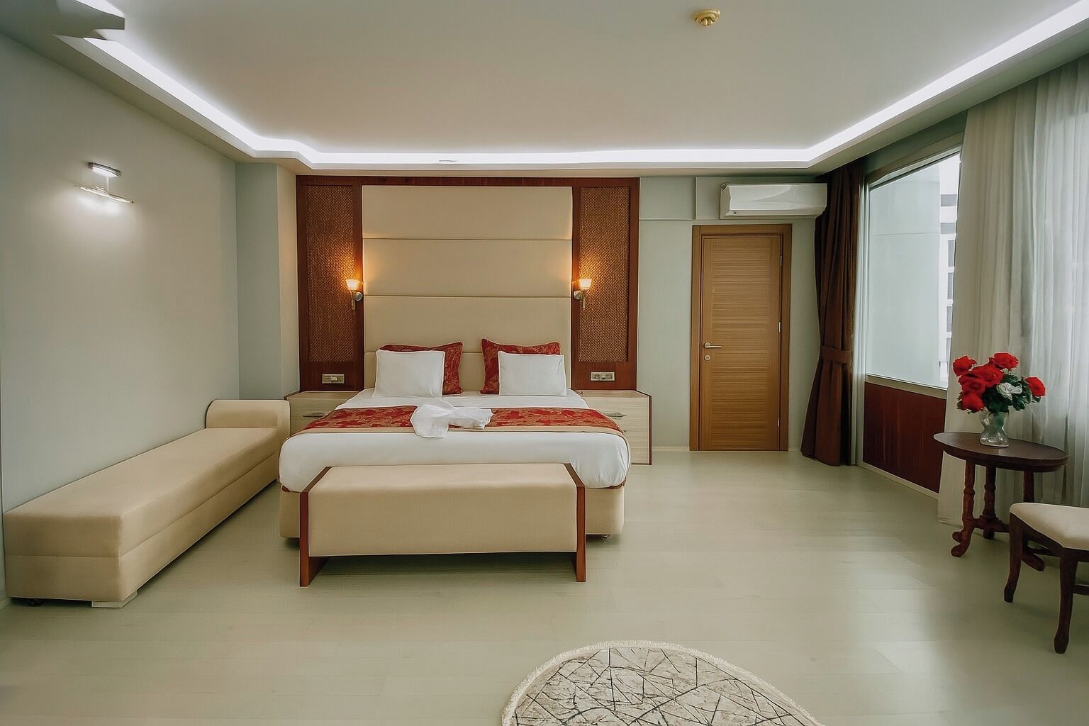 A modern hotel room with a large bed, a sofa, a small table with flowers, and a door.