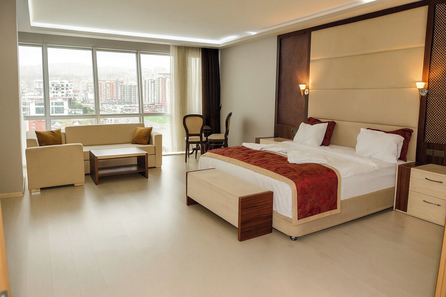 A modern hotel room with a large bed, a sofa, a desk, and a chair. There is a city view through the windows.