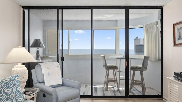 A room with a view of the sea, a chair, a table, and a lamp.