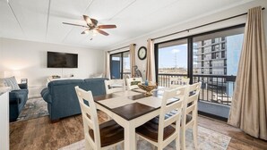 A modern living room with a sofa, dining table, chairs, a ceiling fan, a TV, and large windows with a view of a building and a balcony. - Ocean Princess-Beautiful 2 Bedroom Condo! (Ocean City)