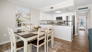 A modern kitchen with white cabinets, stainless steel appliances, and a dining area with a wooden table and chairs. - Ocean Princess-Beautiful 2 Bedroom Condo! (Ocean City)