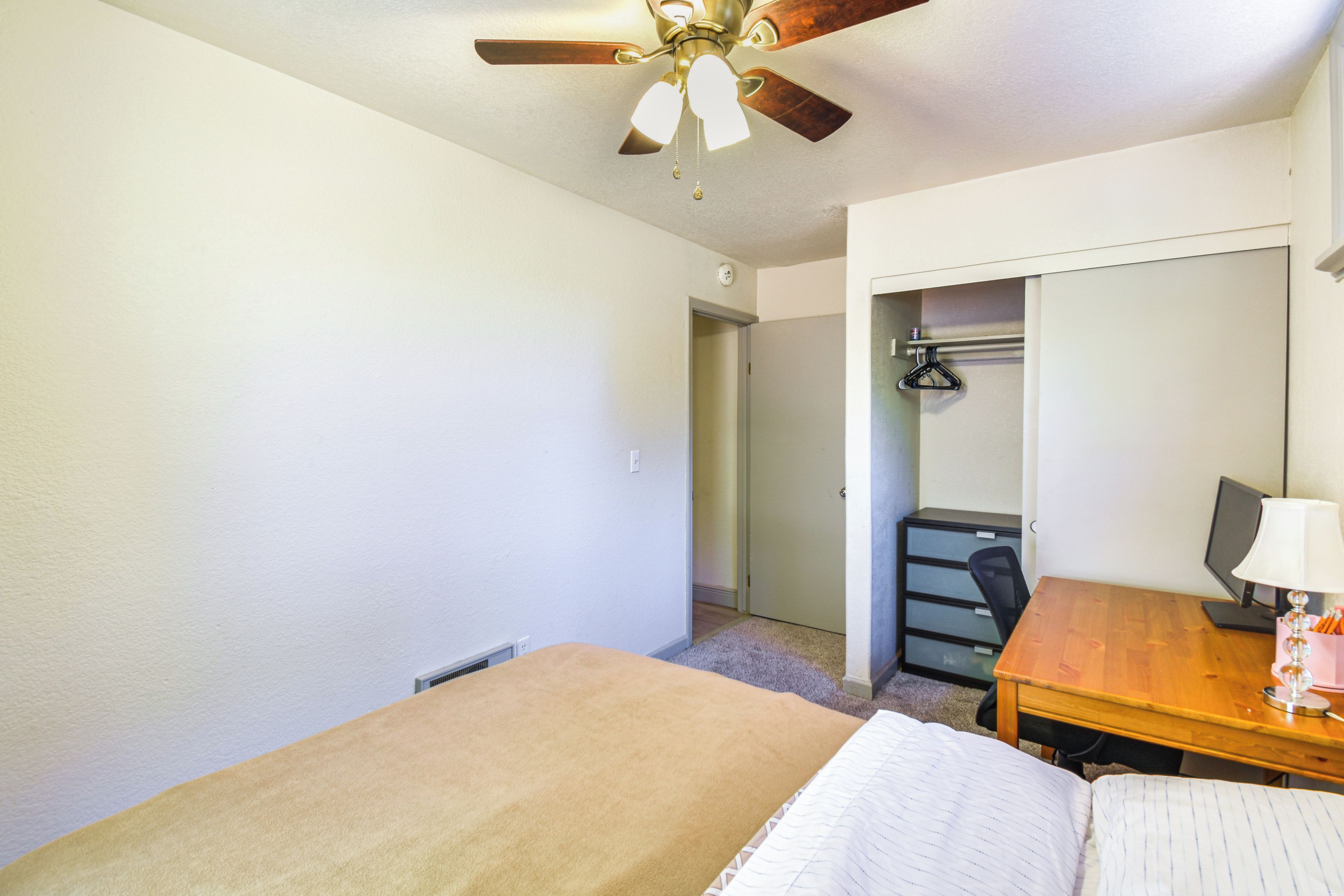 Une chambre à coucher comprenant un lit, un bureau avec un ordinateur, un ventilateur de plafond et une porte donnant sur une salle de bain.