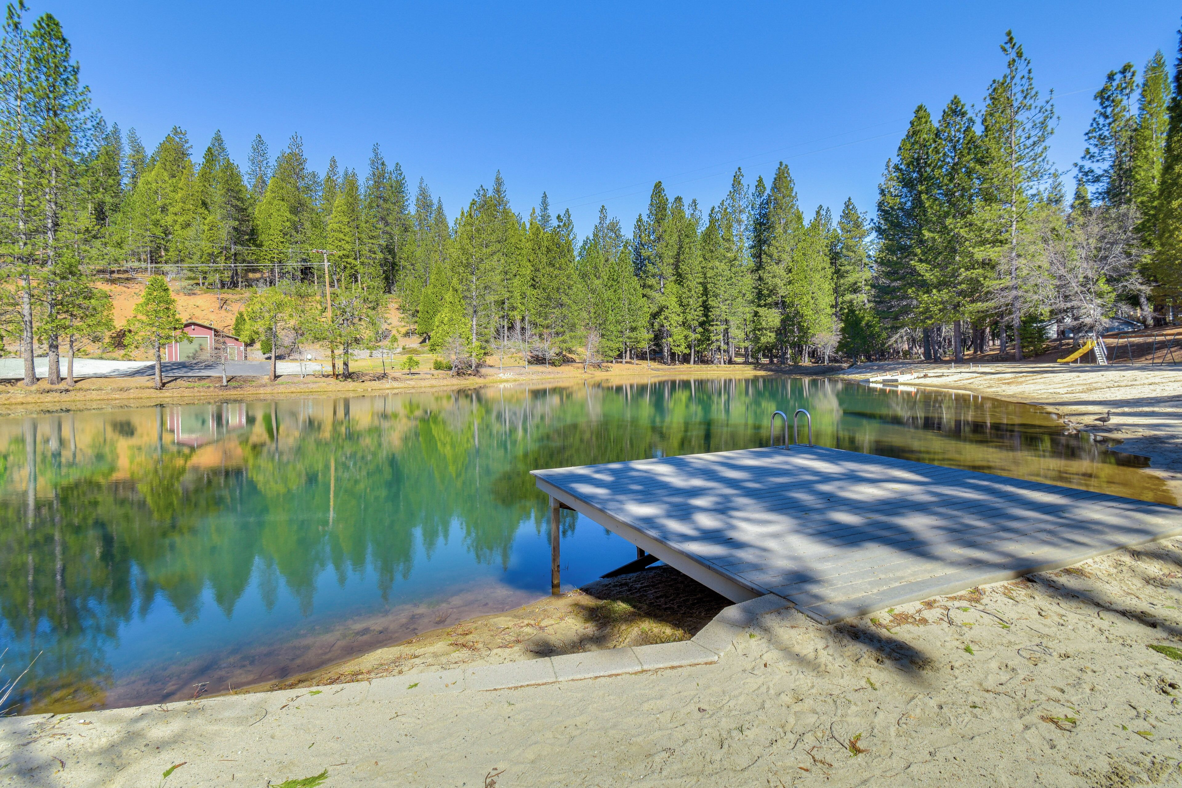A serene outdoor setting with a clear lake, surrounded by trees and a sandy area with a wooden deck.
