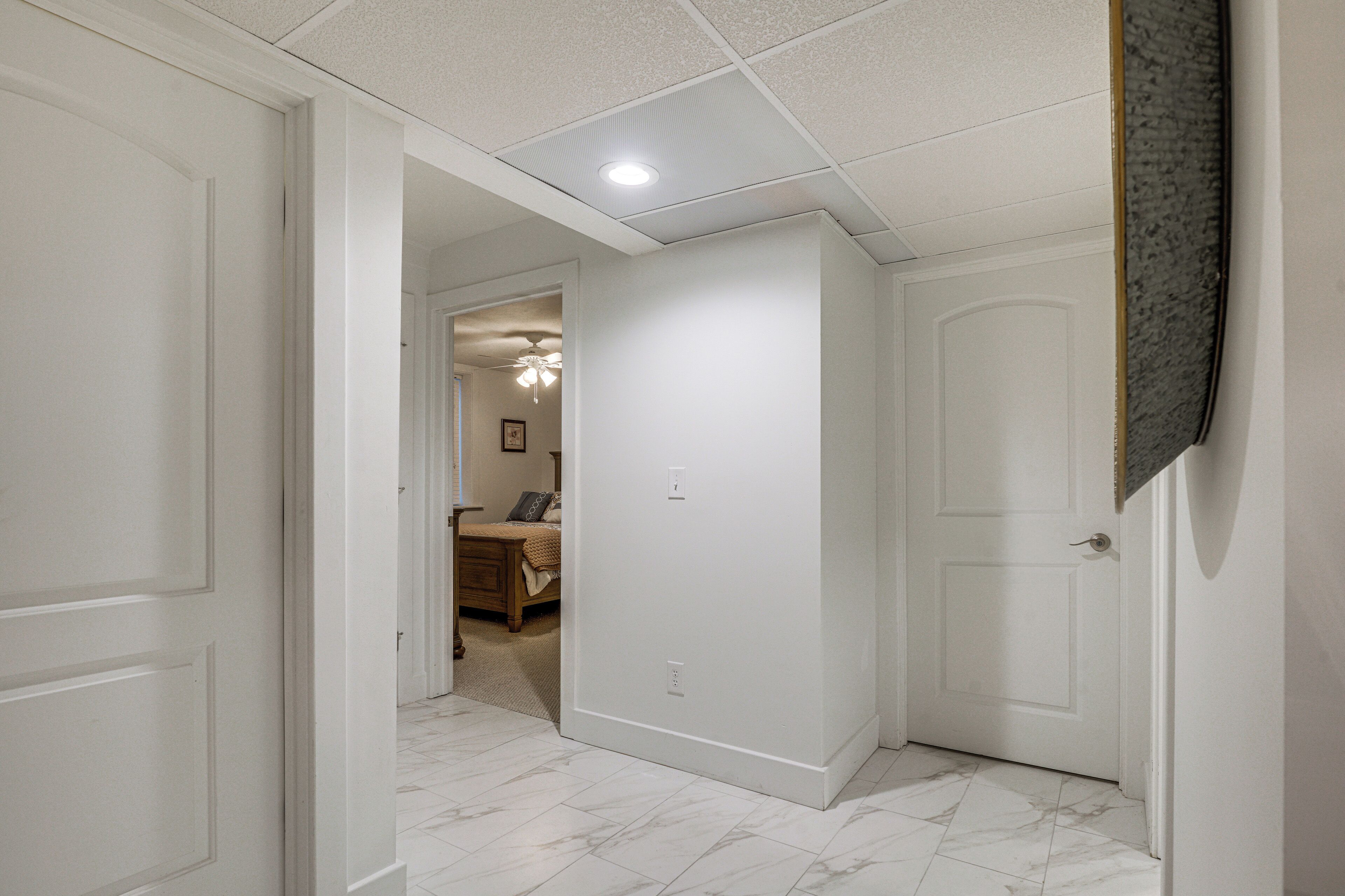 A hallway with white walls and marble flooring, leading to a bedroom with a bed and a ceiling fan.