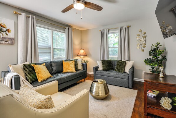 Una sala de estar con un sofá de cuero negro, un sofá gris y un sillón color beige. Hay una lámpara, un jarrón decorativo y una mesita con un libro.