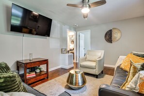 A living room with a flat-screen TV, a ceiling fan, a decorative metal vase, a sofa, a chair, and a small wooden side table with books. - Updated Jamestown Home - 5 Mi to Hpu! (Jamestown)