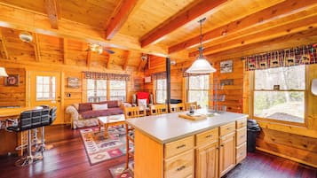 A cozy cabin interior with a wooden interior, a dining area, a kitchen with wooden cabinets, and a large window providing a view of the outdoors.