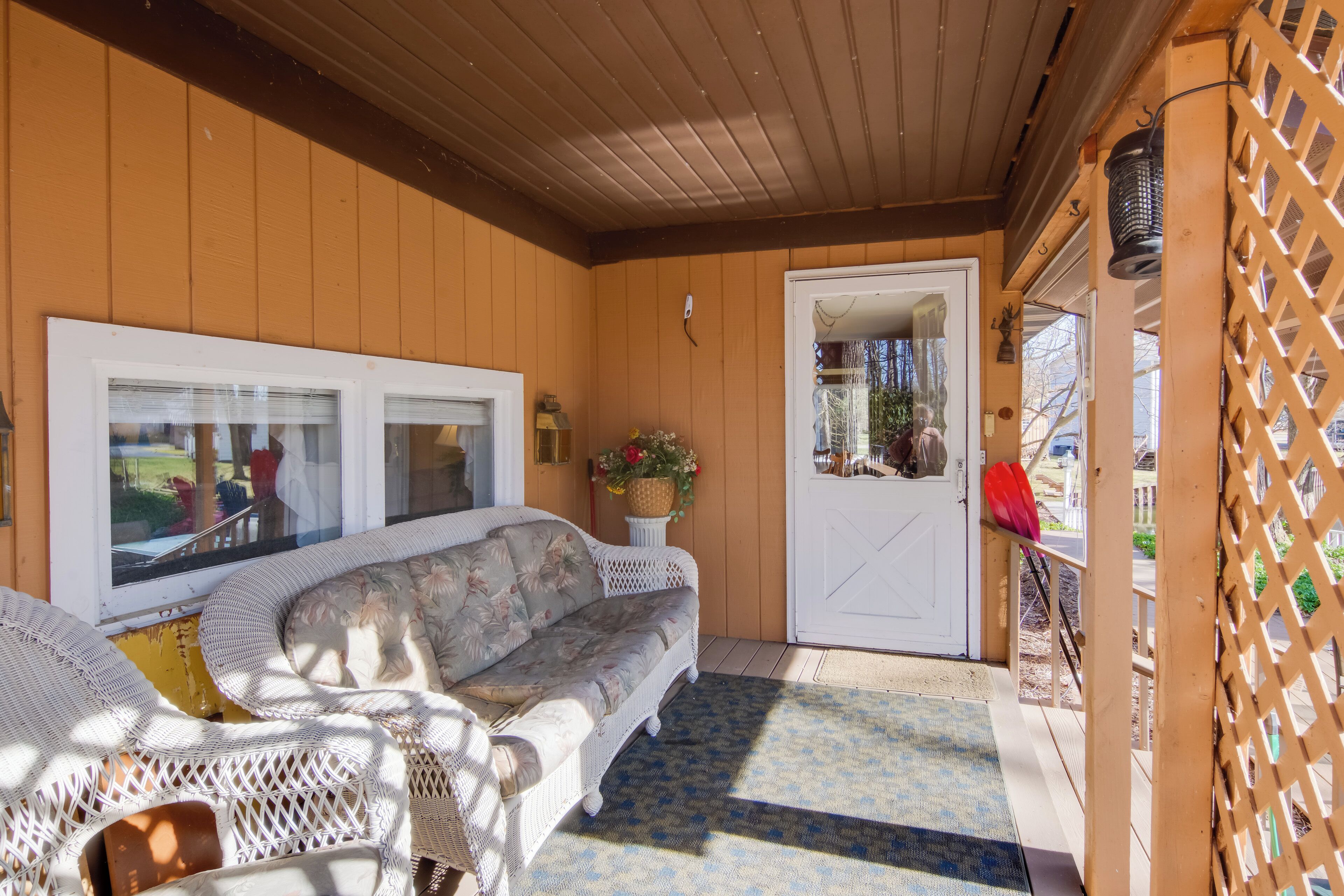 A covered porch with a white wicker sofa, a small table with a basket of flowers, and a wooden lattice wall.