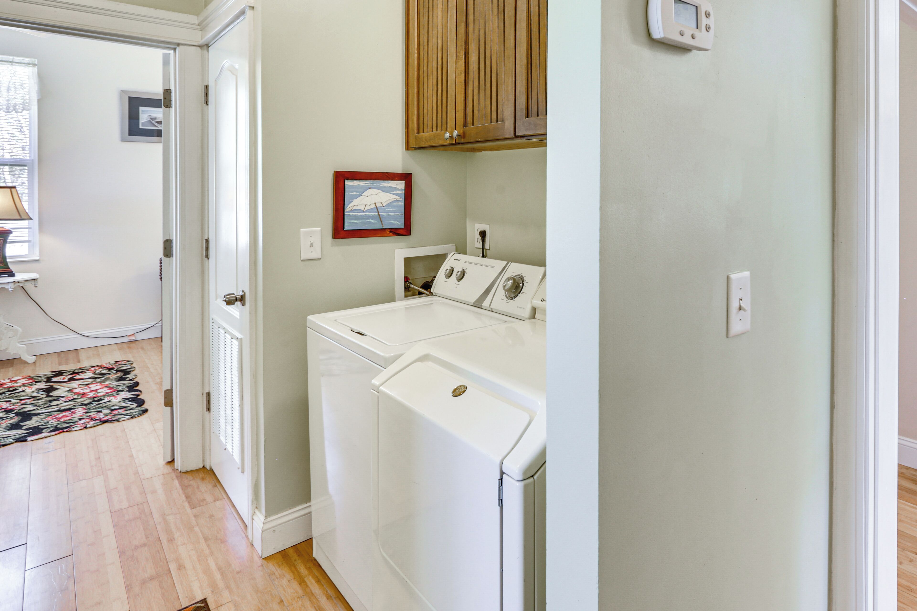 A laundry room with two white washing machines, wooden cabinets above, a power outlet, and a framed picture on the wall.