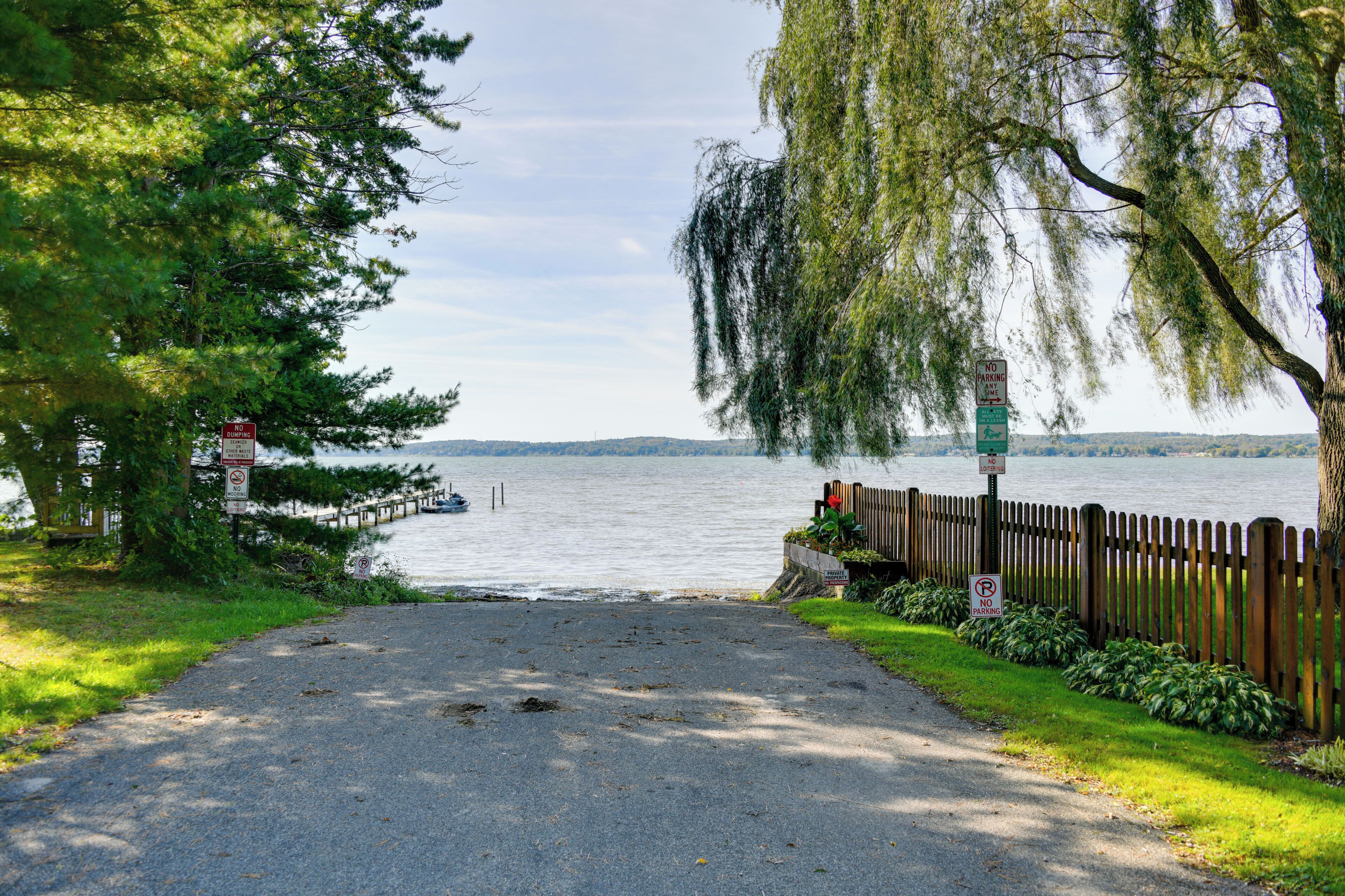 A paved pathway leading to a body of water, flanked by trees and a wooden fence.