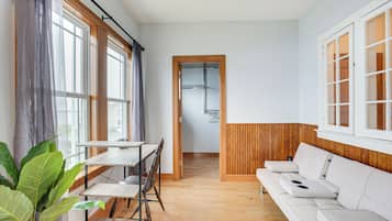 A modern living room with a wooden floor, a white sofa, a dining table with chairs, and large windows with curtains.