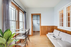 Ein modernes Wohnzimmer mit Holzboden, einem weißen Sofa, einem Esstisch mit Stühlen und großen Fenstern mit Vorhängen.