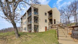 A multi-story building with a stone staircase leading to the entrance, surrounded by a grassy area and trees. - Lake Ozark Condo Rental w/ View & Pool Access! (Lake Ozark)