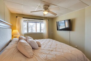 Un dormitorio con cama, ventilador de techo, televisor montado en la pared y ventana con cortinas.