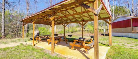 A covered picnic area with wooden benches and picnic tables, surrounded by trees and a grassy field.