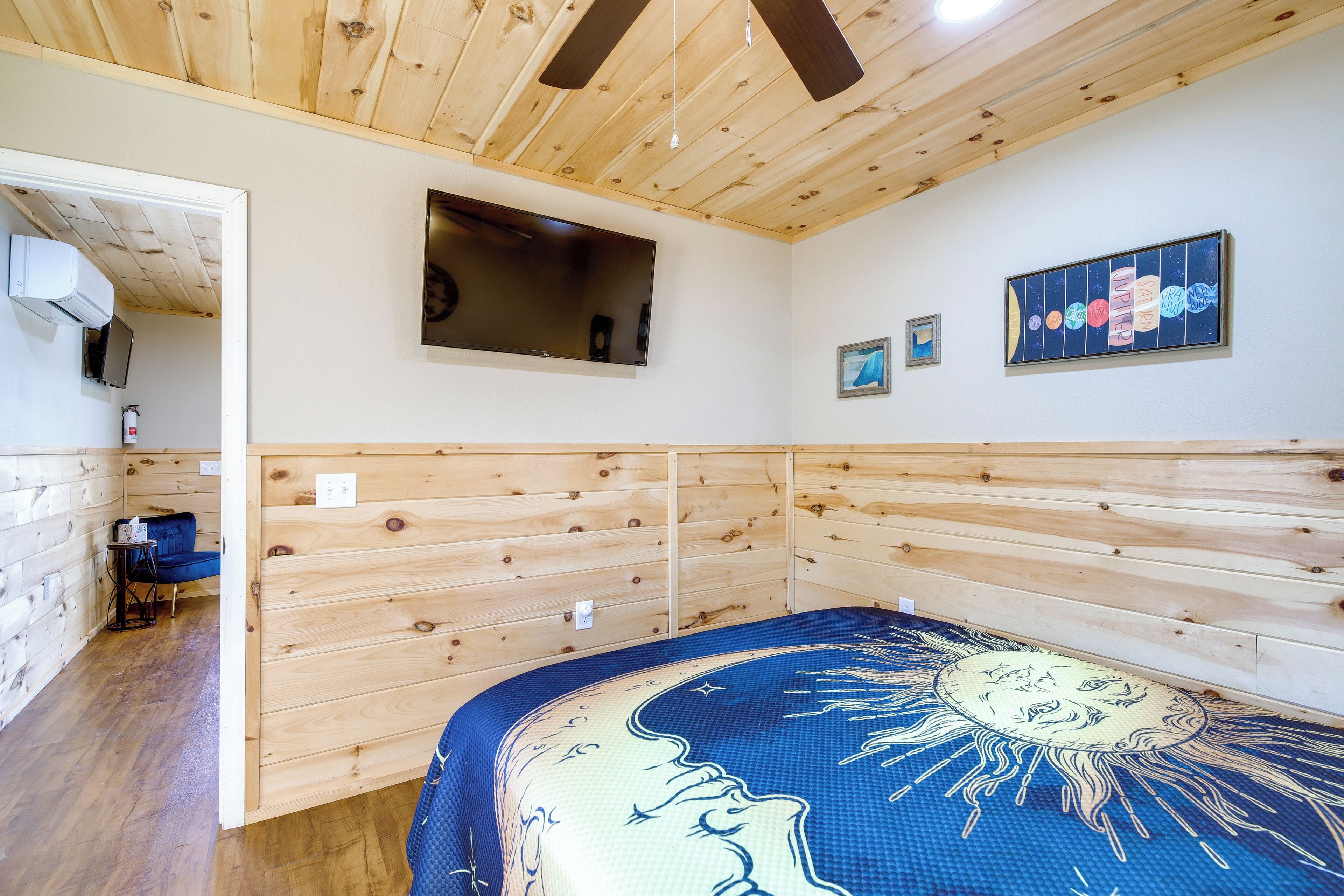 Una habitación con paneles de madera, una cama con colcha azul y blanca estampada, un televisor de pantalla plana montado y un ventilador de techo.