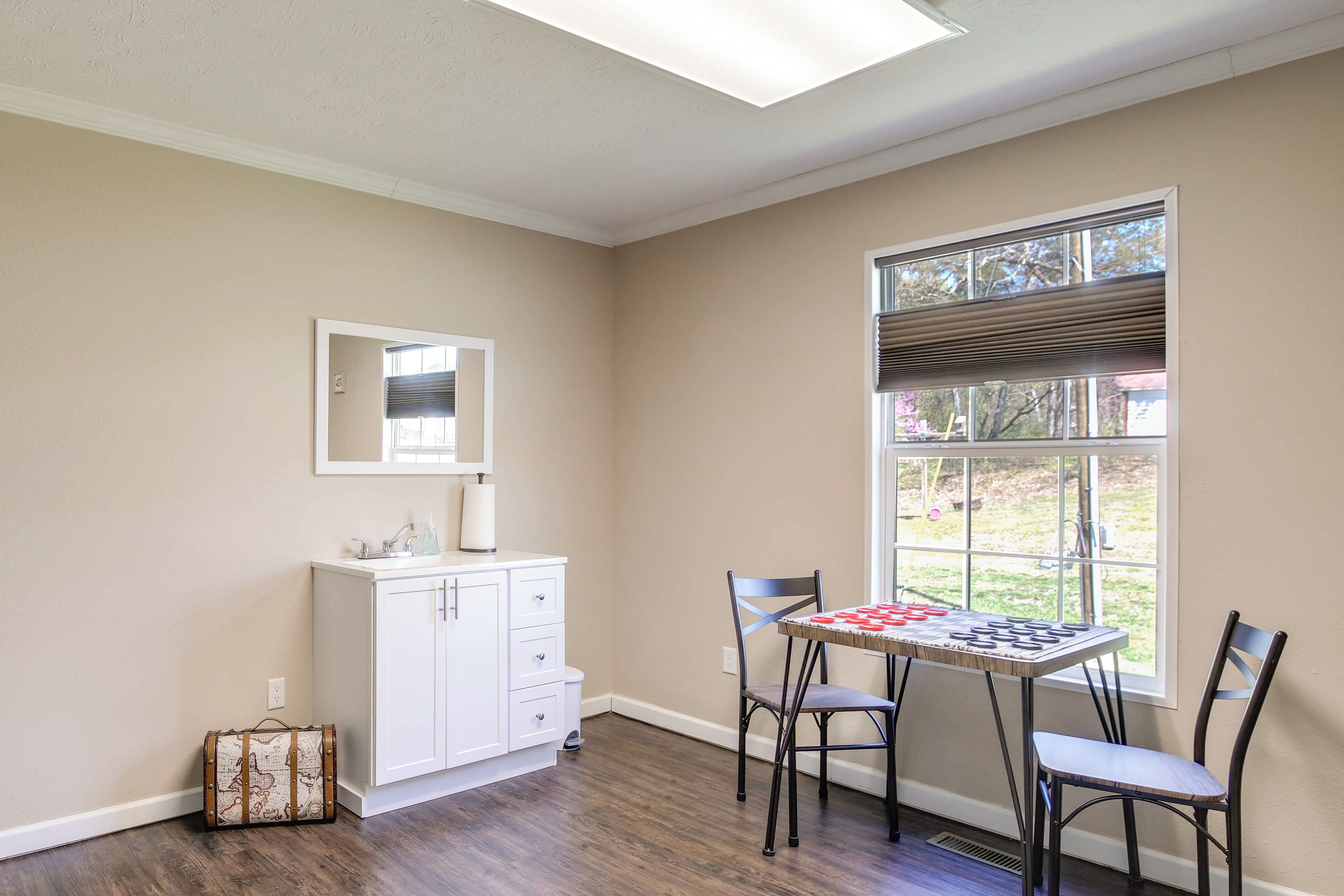 A room with a white cabinet, a table with chairs, a window with blinds, and a suitcase against the wall.