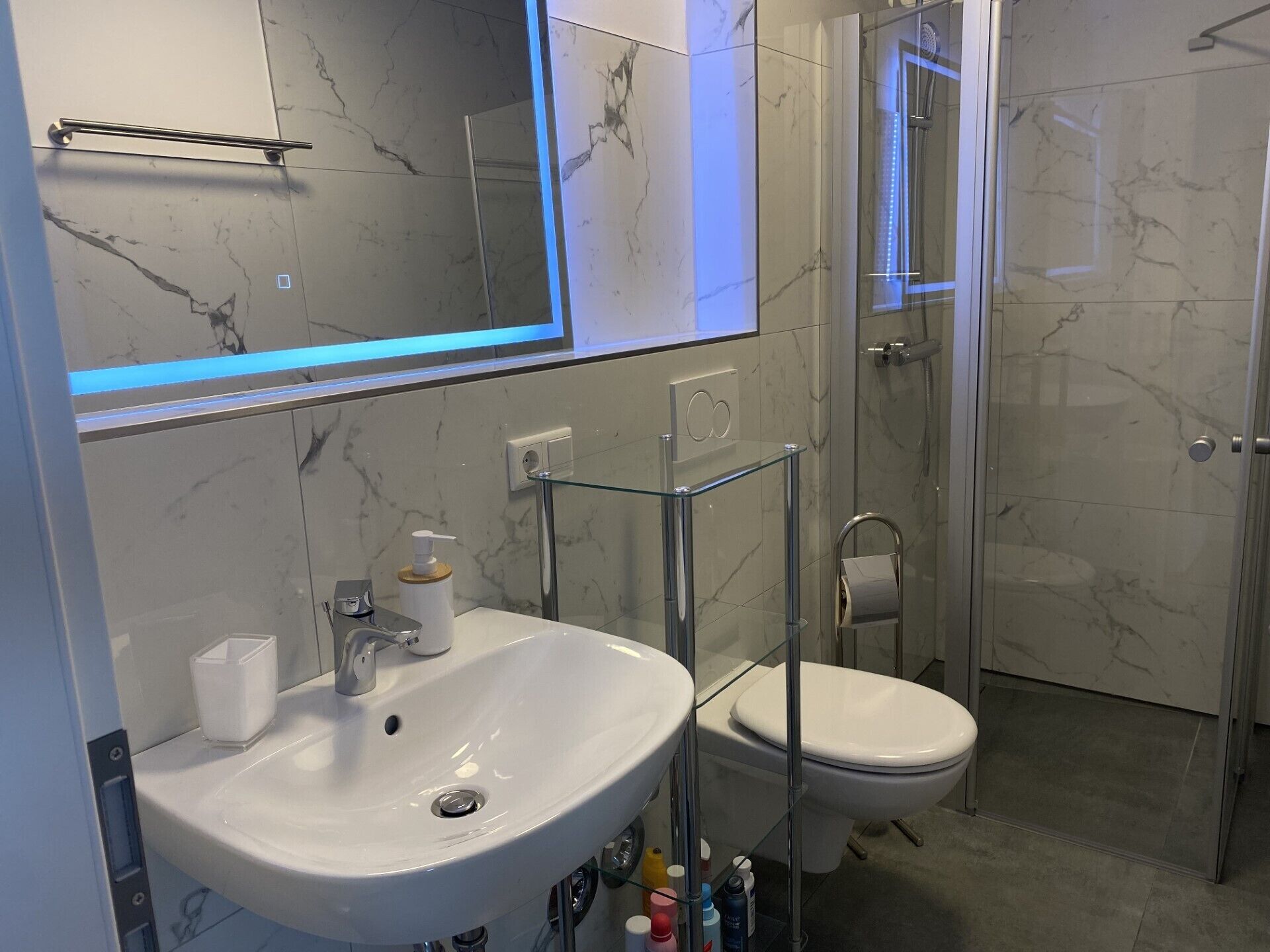 A modern bathroom with a marble-tiled wall, a glass-enclosed shower, a white sink, and a toilet.