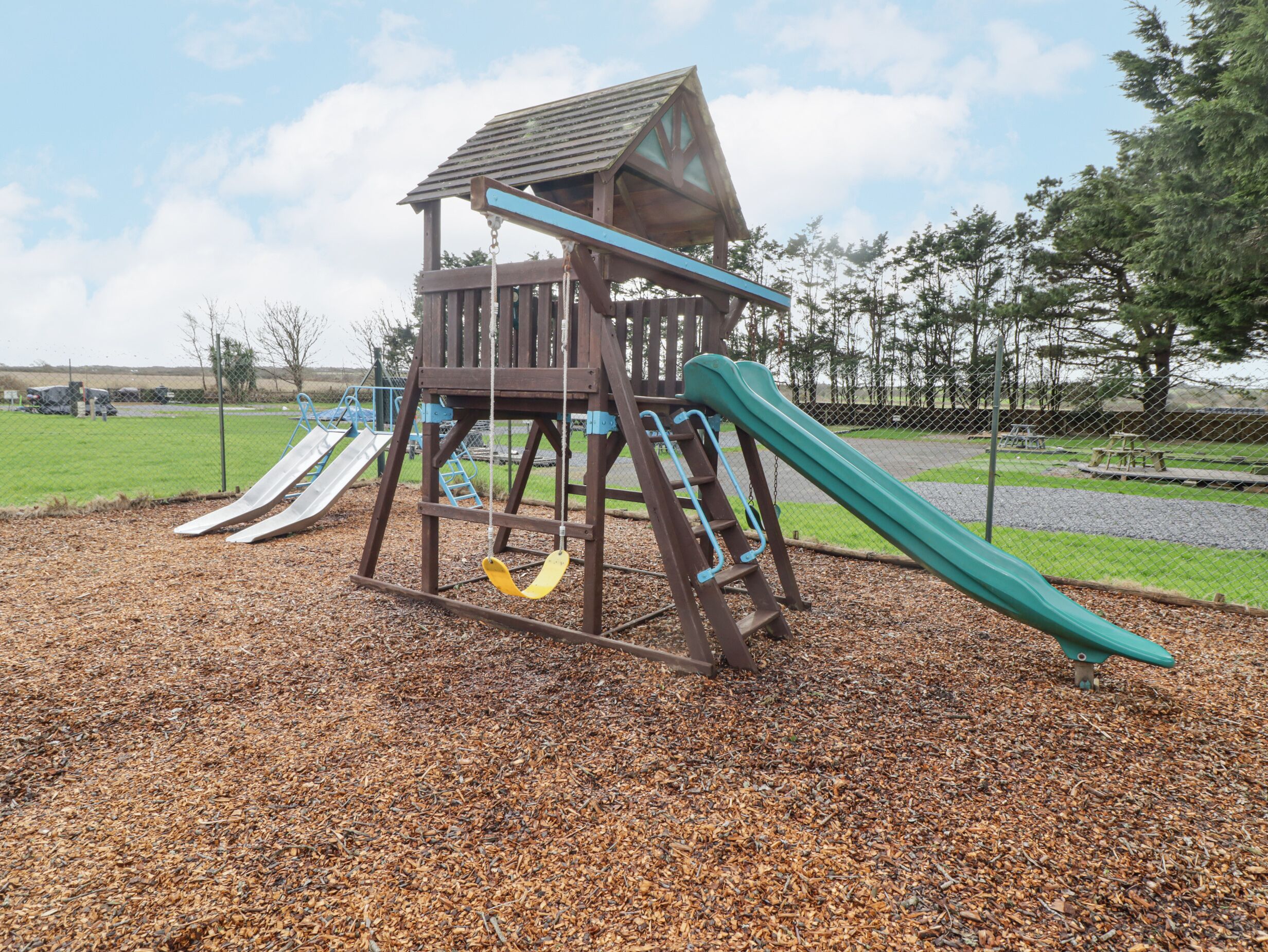 A wooden play structure with a slide and climbing elements, surrounded by a fence and a grassy area.