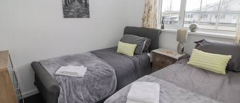 A bedroom with two beds, a nightstand, a lamp, a window with curtains, and a wall-mounted picture.