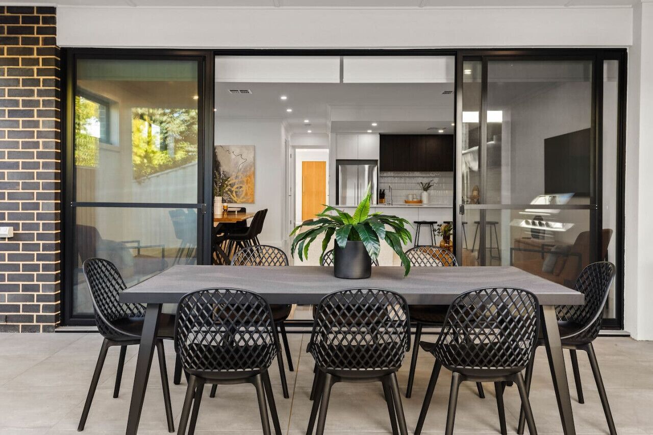 Outdoor dining area with a modern table and several black lattice-patterned chairs. A large potted plant is placed on the table, and the area is adjacent to an indoor space with visible kitchen and living room elements through sliding glass doors. The set
