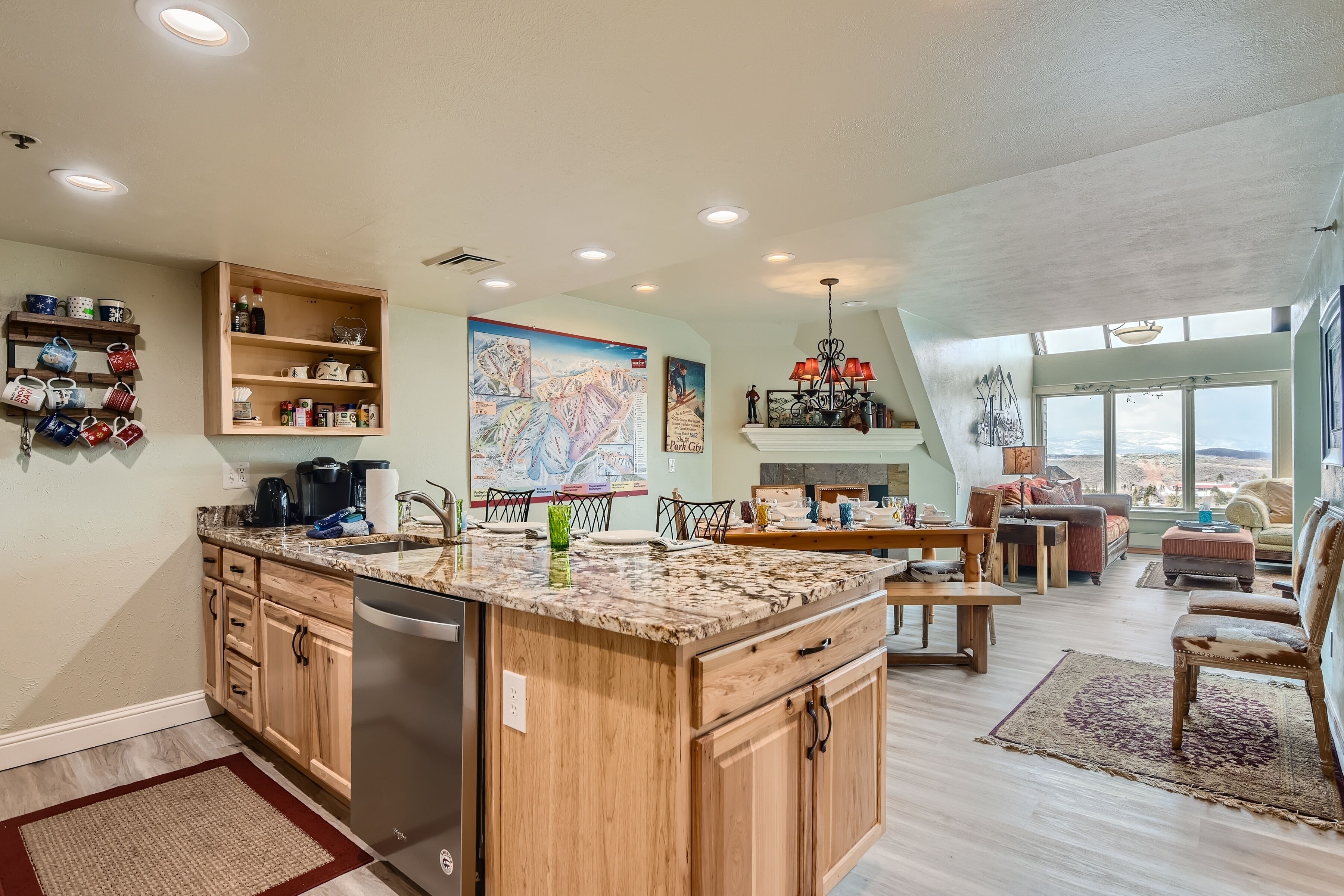A spacious kitchen with wooden cabinets, a granite countertop, and a dishwasher. There is a dining area with a table and chairs, and a large window offering a view of the landscape.