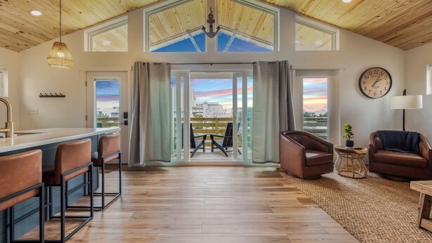 A spacious room with a wooden ceiling, a large window, a bar area with stools, and a seating area with chairs. - OBX Couples Getaway With Hot Tub-Short Walk to the Beach (Rodanthe)