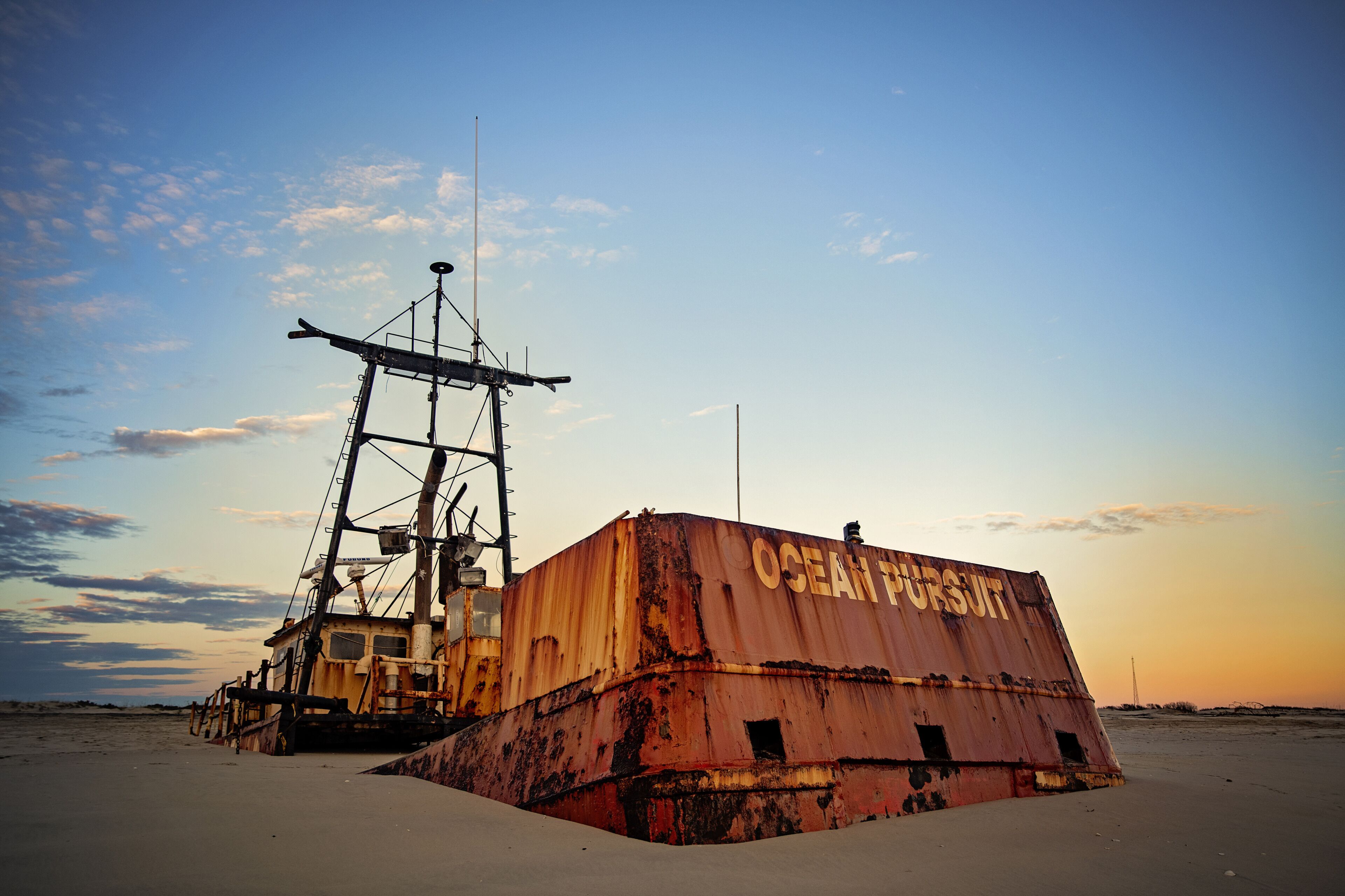 A large, rusted metal structure with the words "OCEAN PURSUIT" on its side, situated on a sandy beach with a clear sky in the background.