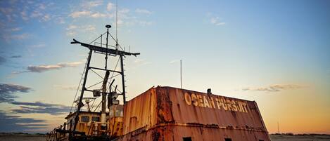 A large, rusted metal structure with the words "OCEAN PURSUIT" on its side, situated on a sandy beach with a clear sky in the background.