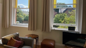 Ein Zimmer mit zwei Fenstern, einem Sofa, einem kleinen Tisch und einem Fernseher.