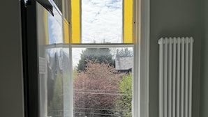 Eine kleine Küche mit einem Edelstahl-Kühlschrank, einer kleinen Mülltonne, einem Fenster mit Blick auf Bäume und ein Haus sowie einem weißen Heizkörper.