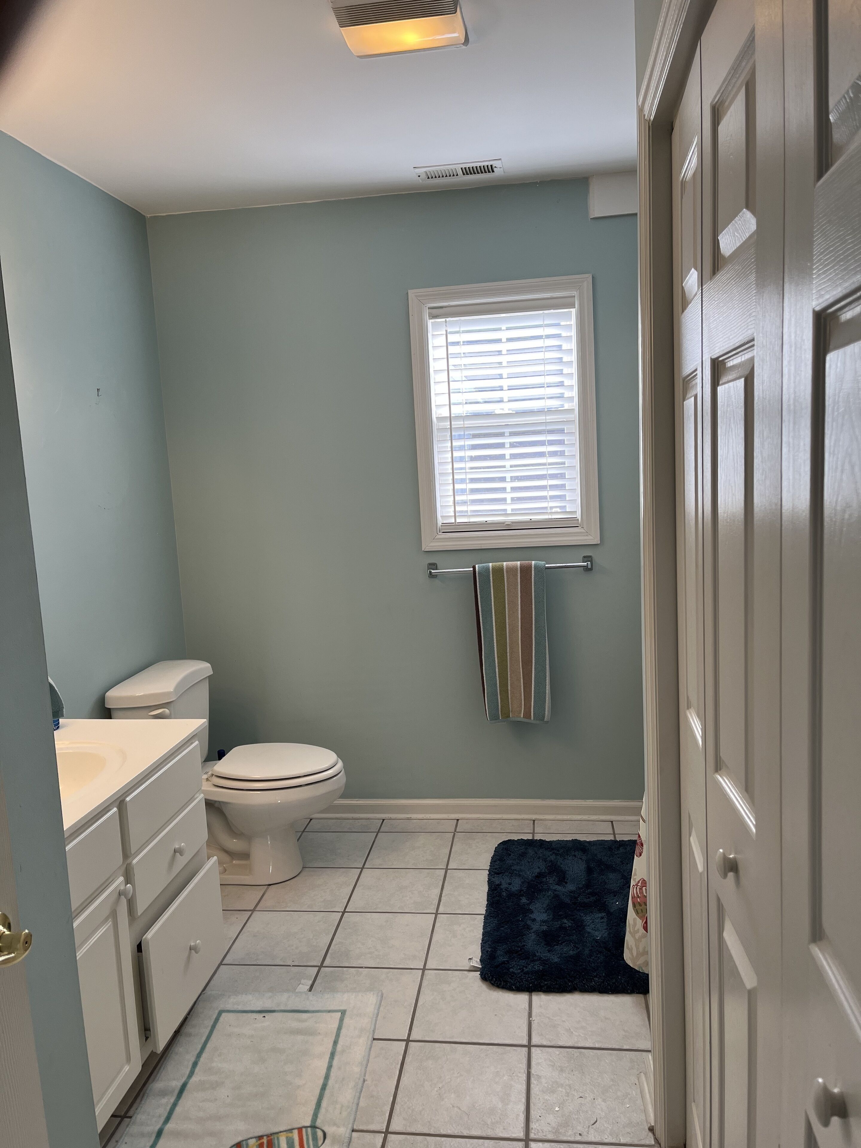 Une salle de bain comprenant des toilettes, un lavabo, une fenêtre avec des stores et un porte-serviettes.