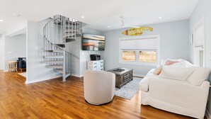 Una sala de estar moderna con una escalera espiral, un televisor de pantalla plana, un sofá blanco y un ventanal con vistas al exterior.