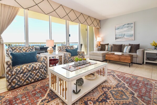 A living room with a patterned sofa, a glass coffee table, and a large window with a view.