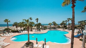 Ein Pool mit gewölbter Kante, umgeben von Liegestühlen und Sonnenschirmen, mit Palmen und Blick auf den Strand.