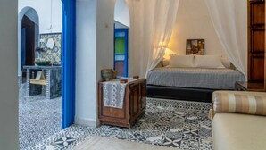 A bedroom with a bed, a nightstand, a small table, and a chair. There is a window with white curtains and a view of an outdoor area with a blue archway. - DarBaali Inn (Rabat)