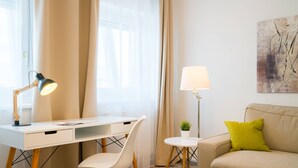 Ett modernt vardagsrum med ett vitt skrivbord, en vit stol, en beige soffa med en grön kudde, en golvlampa och en tavla på väggen.