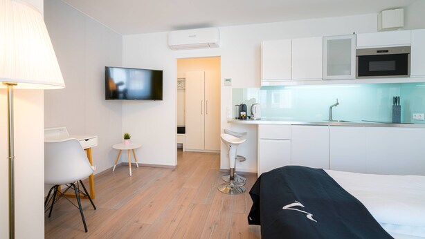 Ett modernt hotellrum med en säng, ett skrivbord, en stol, en liten pall, en TV, ett litet pentry och ett badrum.